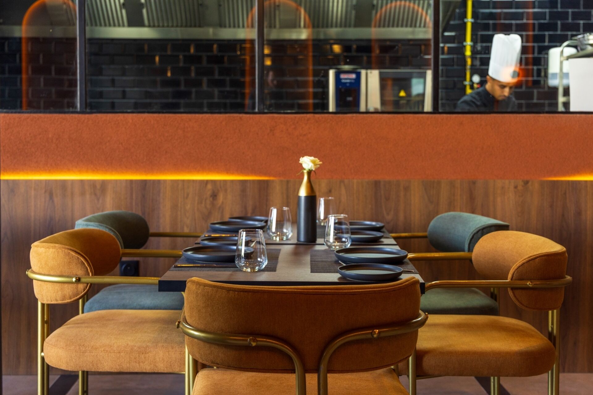 A restaurant with a long table set for dining, chairs with cushioned seats, and a vase with a flower centerpiece.