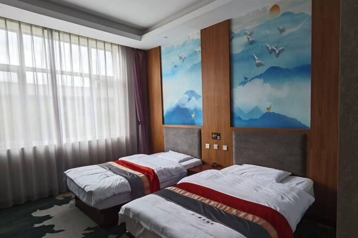 A hotel room with two beds, a large window with sheer curtains, and a wall mural depicting birds and mountains.