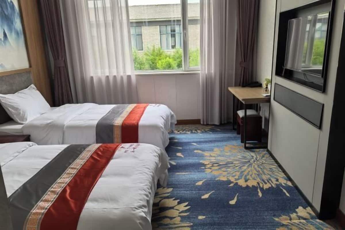 A hotel room with two beds, a desk, a TV, and a window with a view of greenery.