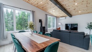 A modern living room with a wooden dining table, green chairs, a grey sofa, a TV, and a large window with a forest view. - Villa Le Frigg | Private Spa • Pet Friendly (Shannon)