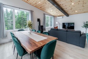 A modern living room with a wooden dining table, green chairs, a grey sofa, a TV, and a large window with a forest view.
