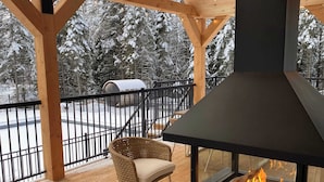 A covered outdoor deck with a wood-burning stove, wicker chairs, and a view of a snowy landscape. - Villa Le Frigg | Private Spa • Pet Friendly (Shannon)