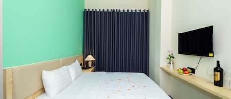 A hotel room with a bed, a nightstand with a lamp, a TV mounted on the wall, and a shelf with fruits and a bottle.