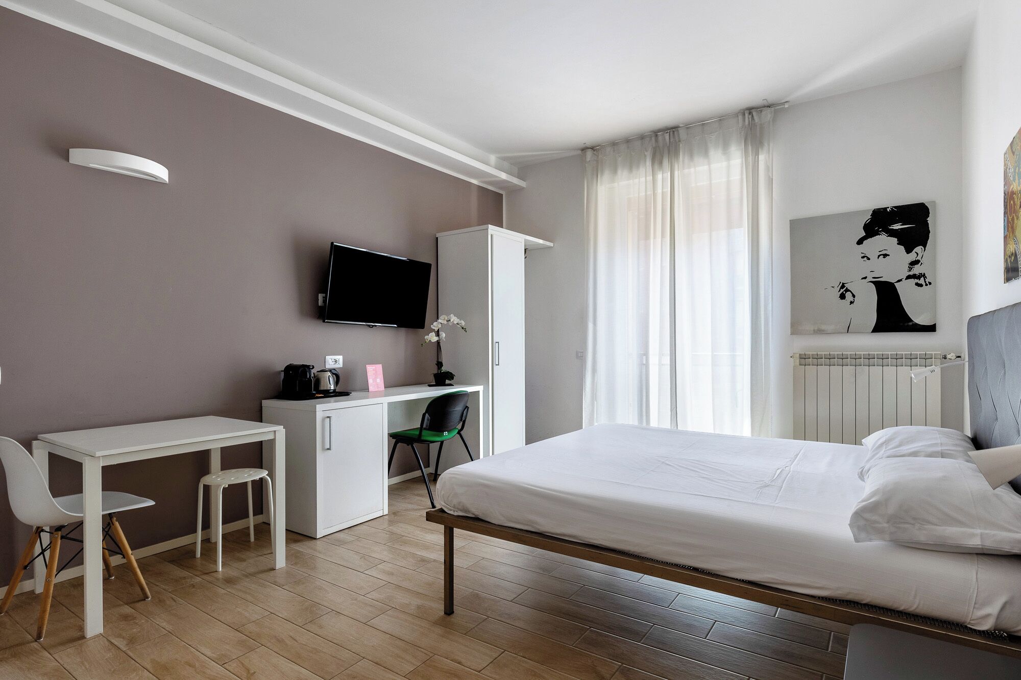Una habitación de hotel con una cama, un escritorio con silla, un televisor, un radiador y un cuadro en la pared.