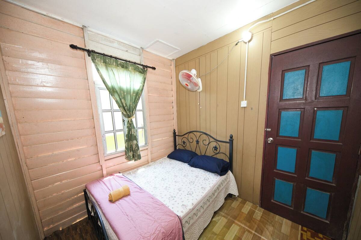 A bedroom with a single bed, a window with a curtain, a wooden door, and a wall-mounted fan.