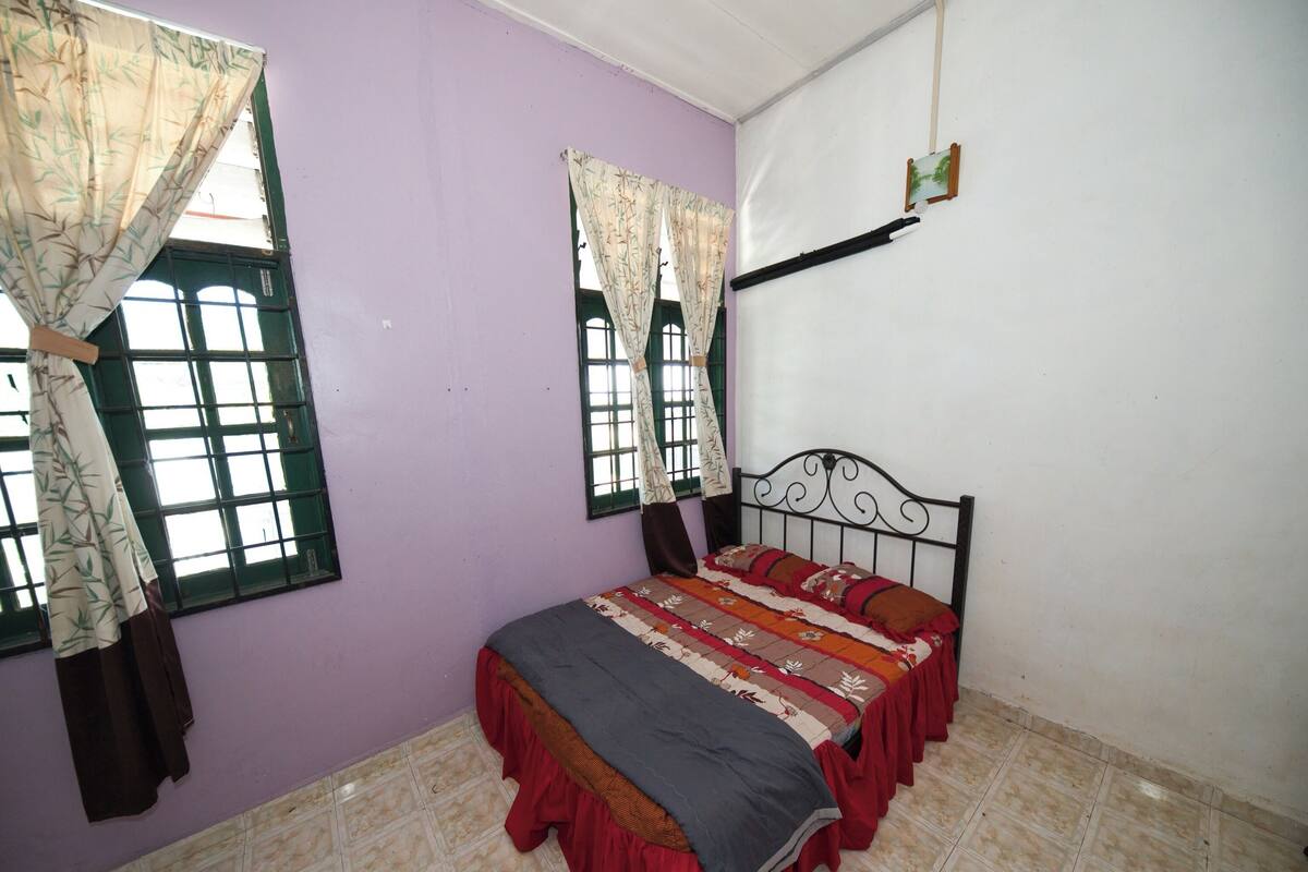 A bedroom with a bed, two windows with curtains, a wall-mounted shelf, and tiled flooring.