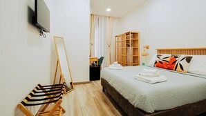 Een netjes ingerichte slaapkamer met een bed, een aan de muur bevestigde televisie, een houten stoel, een boekenkast en een raam met gordijnen.