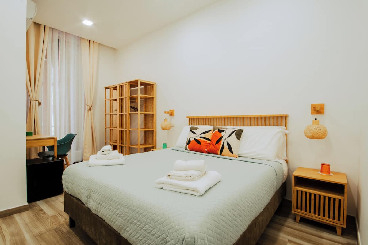 A neatly arranged hotel room with a bed, a wooden wardrobe, a desk with a chair, and a bedside table with a lamp.