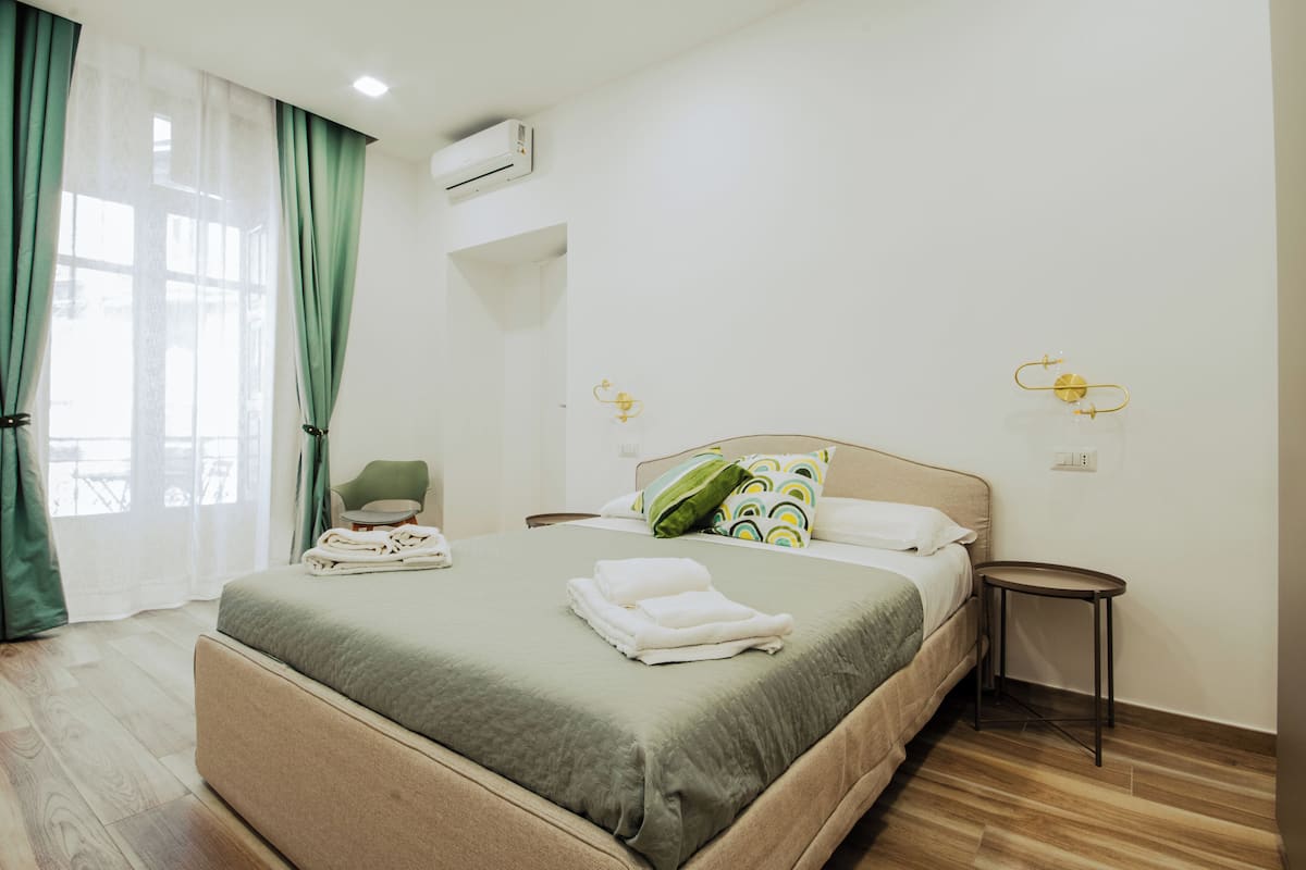 A hotel room with a bed, a chair, a small table, and a window with curtains.