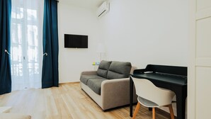 Een moderne woonkamer met een bank, een bureau met stoel, een flatscreen-televisie en een raam met gordijnen.