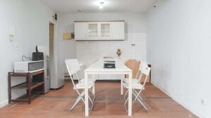 A compact kitchen with white cabinets, a white table, and two white chairs. A microwave is on a wooden shelf, and a small appliance is on the floor. - Apartemen Taman Rasuna 2BR. 80sqm (Jakarta)