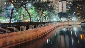 A nighttime view of high-rise buildings with illuminated windows, a reflective water body, and trees in the foreground. - Apartemen Taman Rasuna 2BR. 80sqm (Jakarta)