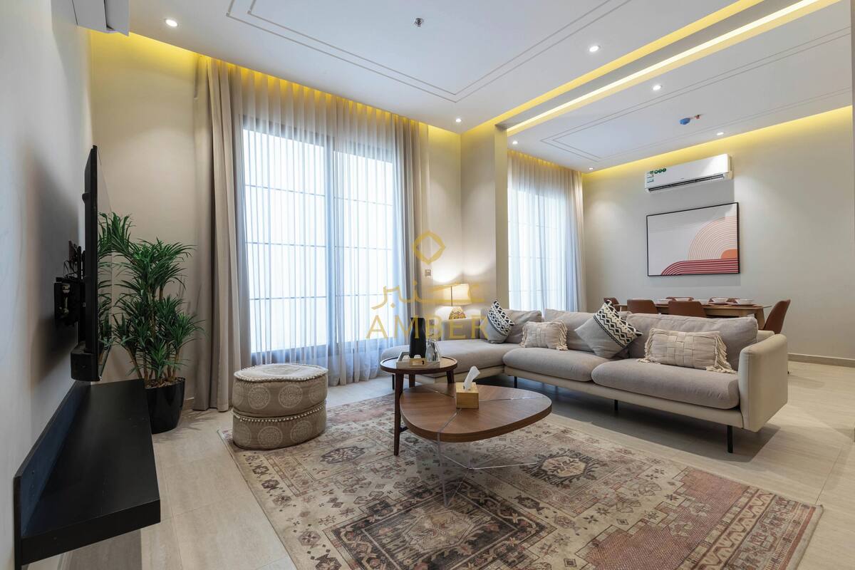A modern living room with a sofa, coffee table, and a TV. There are curtains, a plant, and a rug.