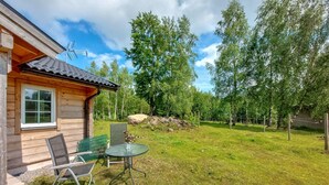 Puuinen mökki, jonka pihalla on metallinen pöytä ja tuoleja. Mökkiä ympäröivät puut, ja taivas on kirkas.