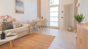 A modern living room with a white sofa, a dining table with chairs, a small side table with a vase and magazine, and a large window with a view of the outdoors. - The Printers Home in Como (Como)