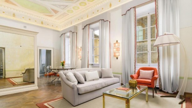 Un salon spacieux avec un canapé gris, un fauteuil rouge et une table basse en verre. On aperçoit à l’arrière-plan un coin repas avec une table et des chaises.