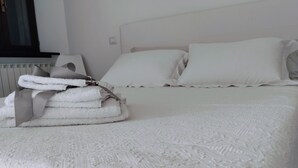 En pænt redt seng med hvide sengetøj og puder, et foldet håndklæde ved siden af, og en radiator under vinduet.