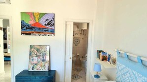 A bedroom with a bed, a painting, a blue chest of drawers, a nightstand, and a bathroom visible through an open door. - Casa FeFe a scenic view of Positano and a swimming pool! (Positano)