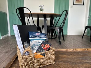 Un panier en osier contenant un livre, un sachet de sel marin et un paquet de café, posé sur une table en bois dans un coin repas avec des chaises noires et une porte verte.