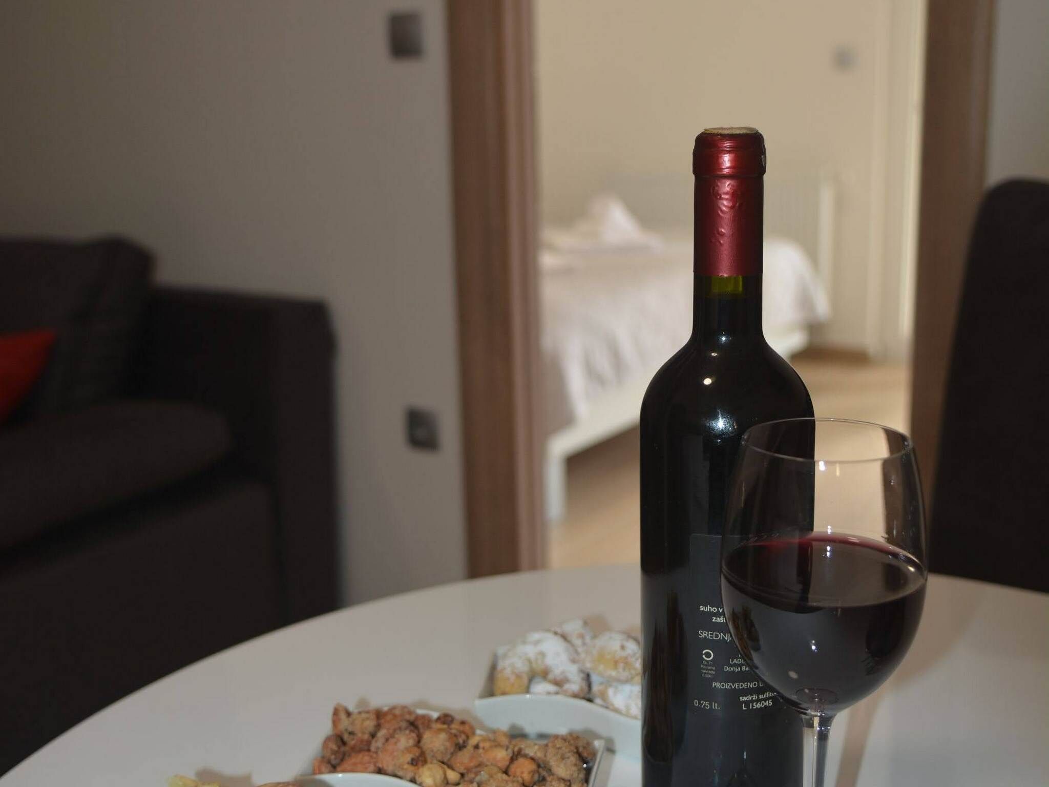 A bottle of red wine and a glass on a table, with a plate of nuts and a small bowl of what appears to be a dessert or snack.