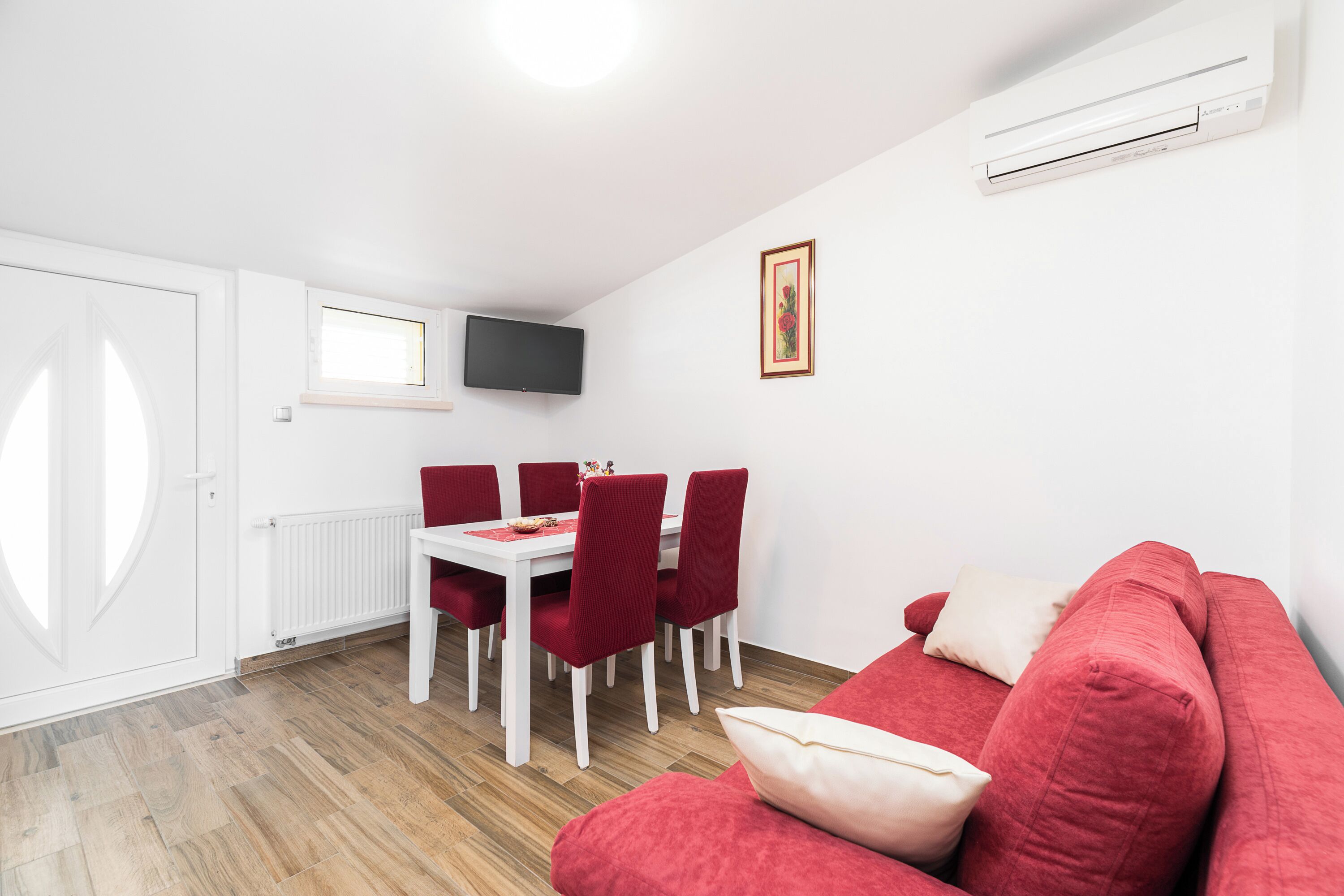 A modern living room with a red sofa, a dining table with red chairs, a flat-screen TV mounted on the wall, and a white radiator.