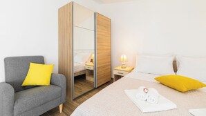 A hotel room with a grey sofa, a bed with white and yellow pillows, a wooden wardrobe, a bedside table with a lamp, and a towel on the bed. - Fancy Apartments Dream (Smokvica)