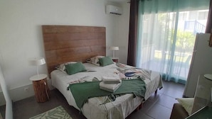 Een slaapkamer met een houten hoofdbord, twee bedden, een tapijt met groene en witte patronen, een kleine ronde tafel met een lamp en uitzicht op een balkon door dunne gordijnen.