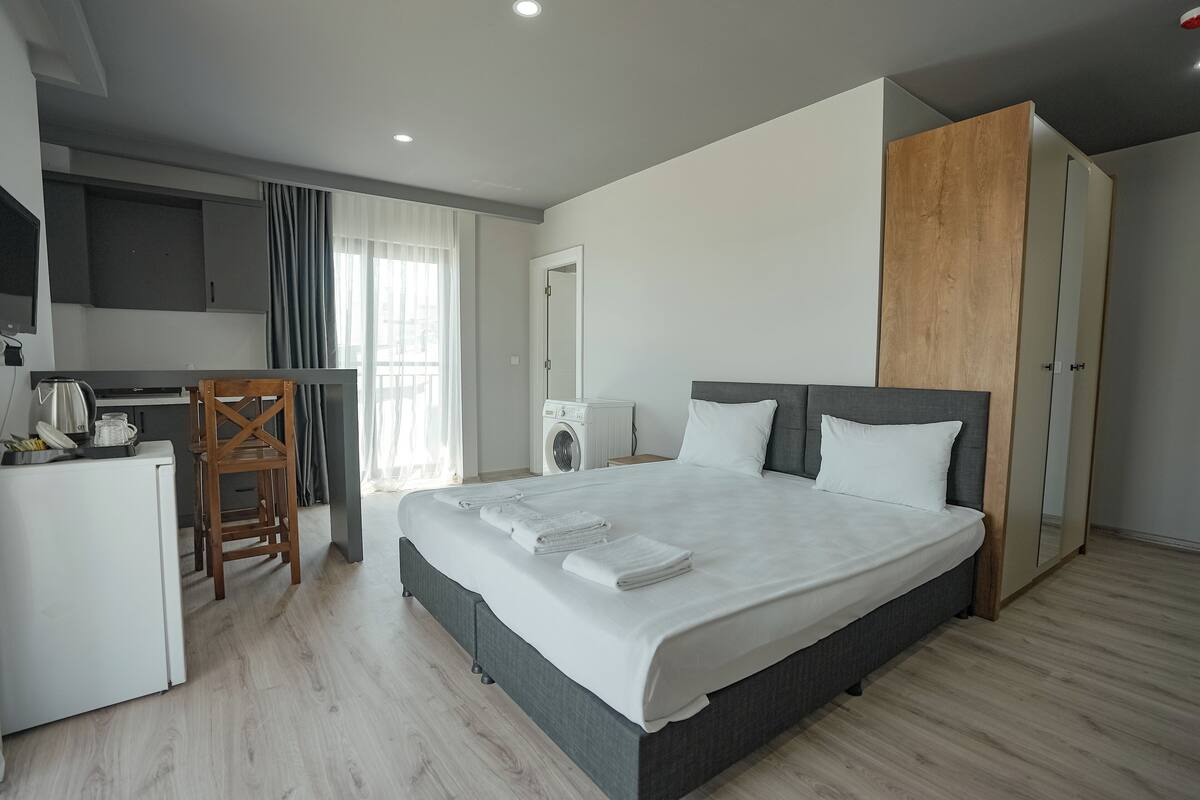 A modern hotel room with a large bed, a desk with a kettle, a washing machine, and a wardrobe.