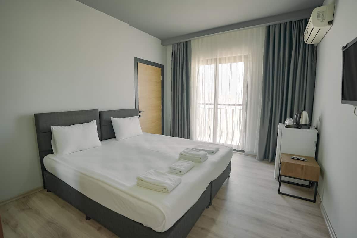 A neatly made bed with white linens and towels, a bedside table with a remote control, and a window with curtains.