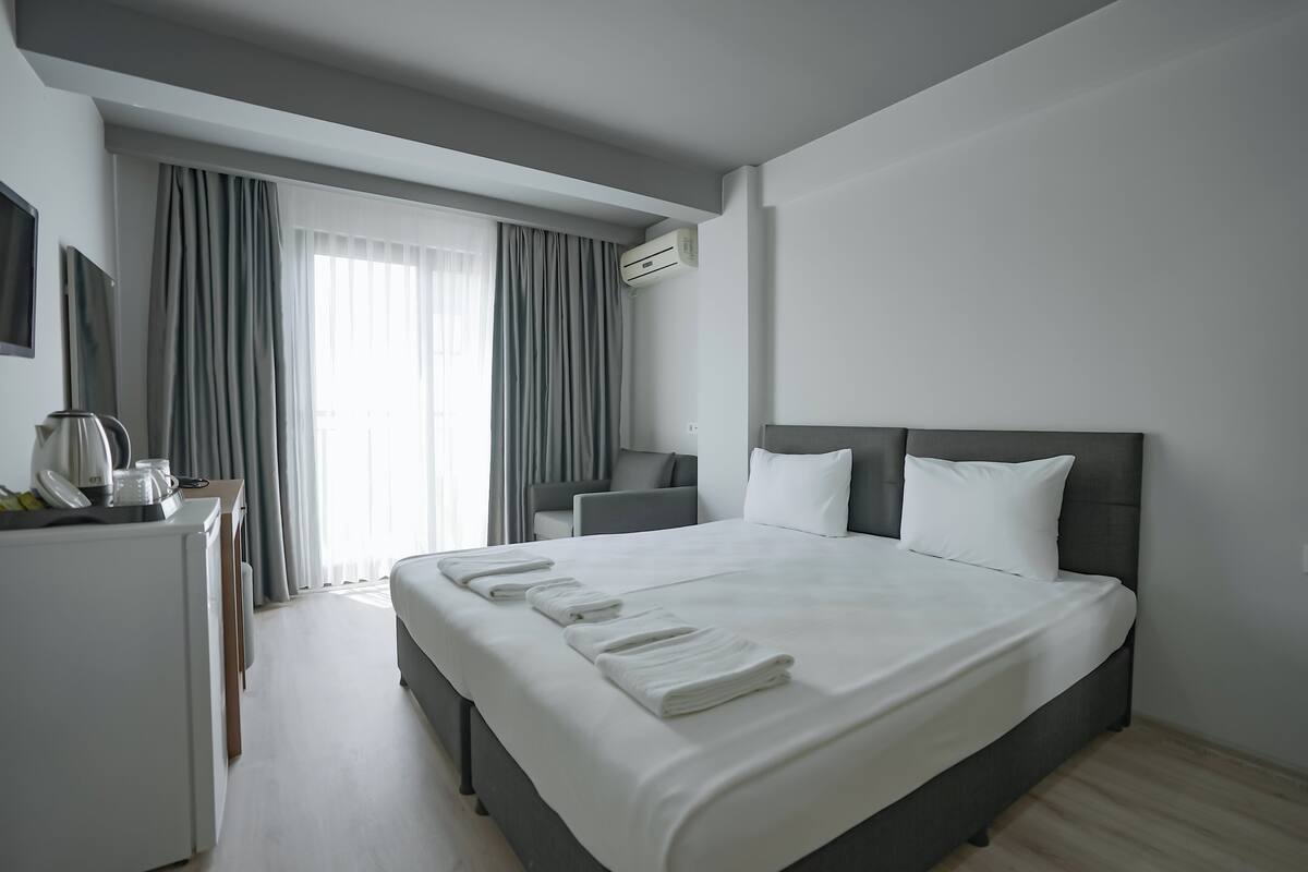 A modern hotel room with a large bed, a desk with a kettle, and a view through sheer curtains.