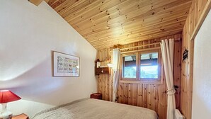 A bedroom with a wooden ceiling, a bed with a beige blanket, a bedside table with a lamp, a framed picture on the wall, and a window with curtains. - Modern Apartment with Mountain Views, Near Combloux Center (Combloux)