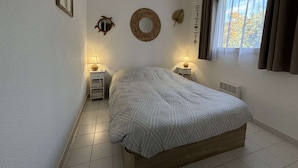 A bedroom with a bed, two bedside tables with lamps, a window with curtains, and wall decorations including a mirror and a circular woven item.