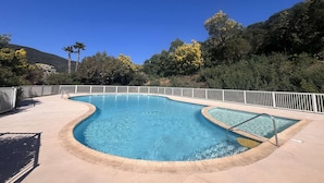 A swimming pool with a curved edge, surrounded by a white fence, set against a backdrop of trees and a clear blue sky.