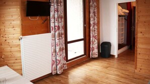 A room with wooden walls, a radiator, a TV mounted on the wall, a window with patterned curtains, a trash bin, and a door leading to another room. - Studio for 2 in the heart of Les Orres 1650 with balcony and ski locker (Les Orres)