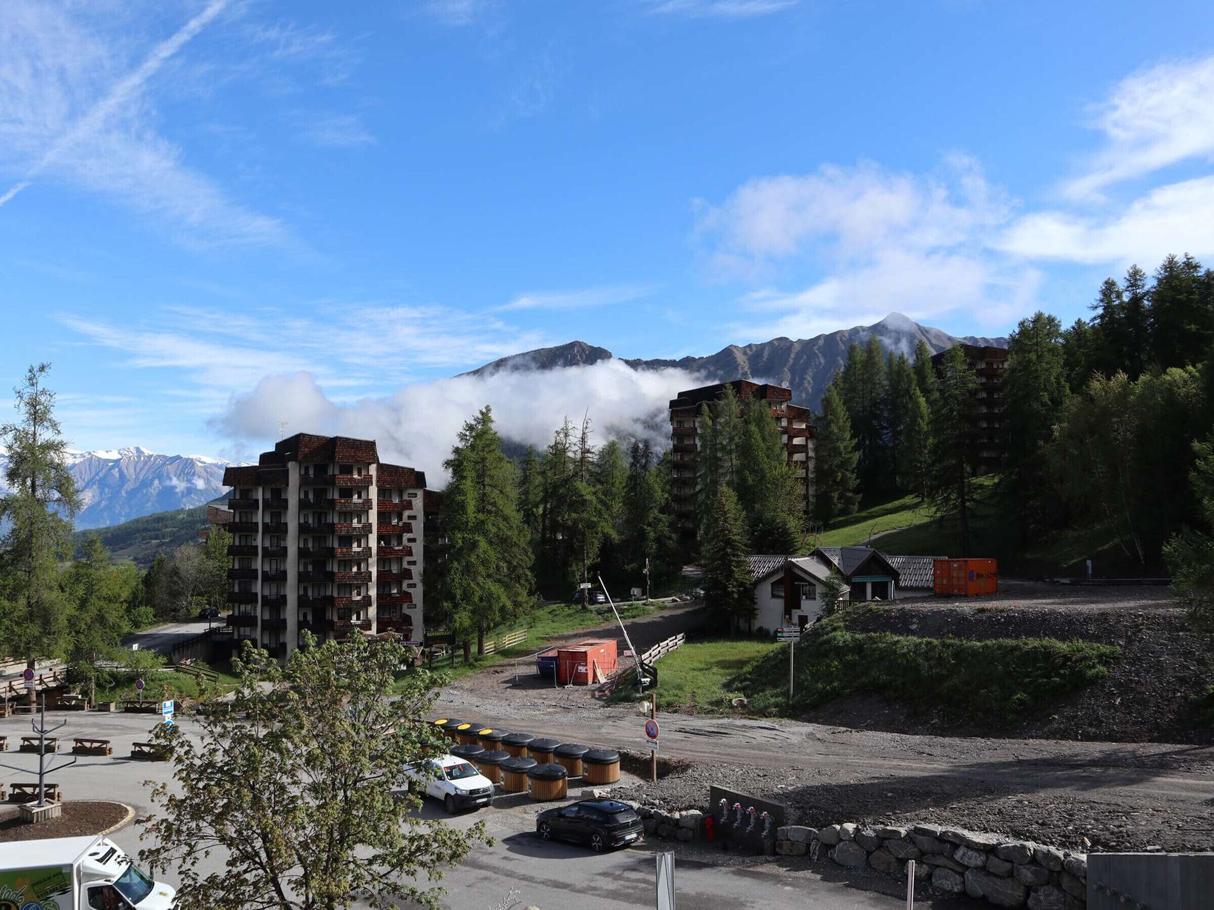 Gebirgslandschaft mit Parkplatz, einem mehrstöckigen Gebäude und Blick auf schneebedeckte Berge.
