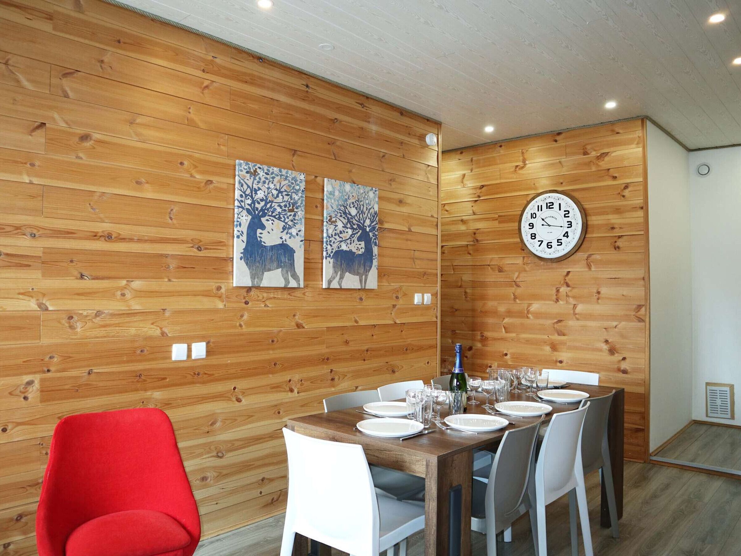 A dining area with a wooden wall, two paintings, a clock, and a table set for six.