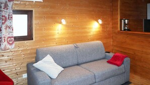 A grey sofa with a white pillow and a red blanket, wooden walls, a window with a curtain, and a coffee machine on a shelf. - Cozy studio for 2 in the heart of Les Orres 1650 (Les Orres)