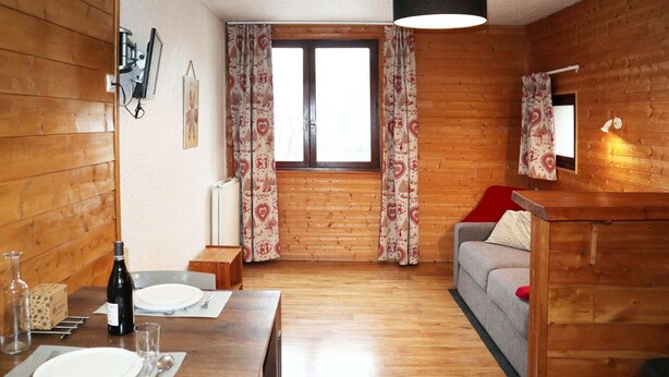 A cozy cabin-style room with wooden walls, a dining area with a table set for two, a gray sofa, and a window with patterned curtains. - Cozy studio for 2 in the heart of Les Orres 1650 (Les Orres)