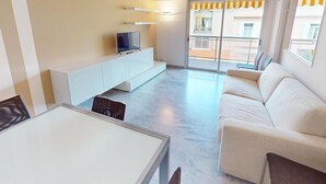 Una sala de estar moderna con un sofá, un televisor sobre un mueble blanco, una mesa de comedor y un balcón con toldos a rayas amarillas.
