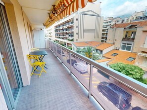 En balkon med et bord og stole, med udsigt over en gade med parkerede biler og bygninger.