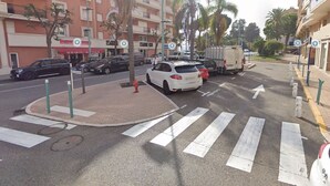 Vista de la calle con coches aparcados al borde, un paso de peatones y edificios con balcones.