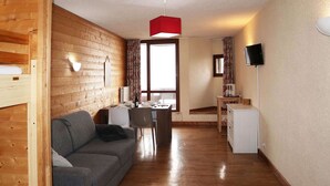 A cozy room with a wooden wall, a gray sofa, a dining table with chairs, a small kitchenette, and a TV mounted on the wall.