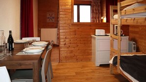 A cozy cabin with a wooden dining table set for four, a bunk bed, a small kitchenette, and a window with curtains.