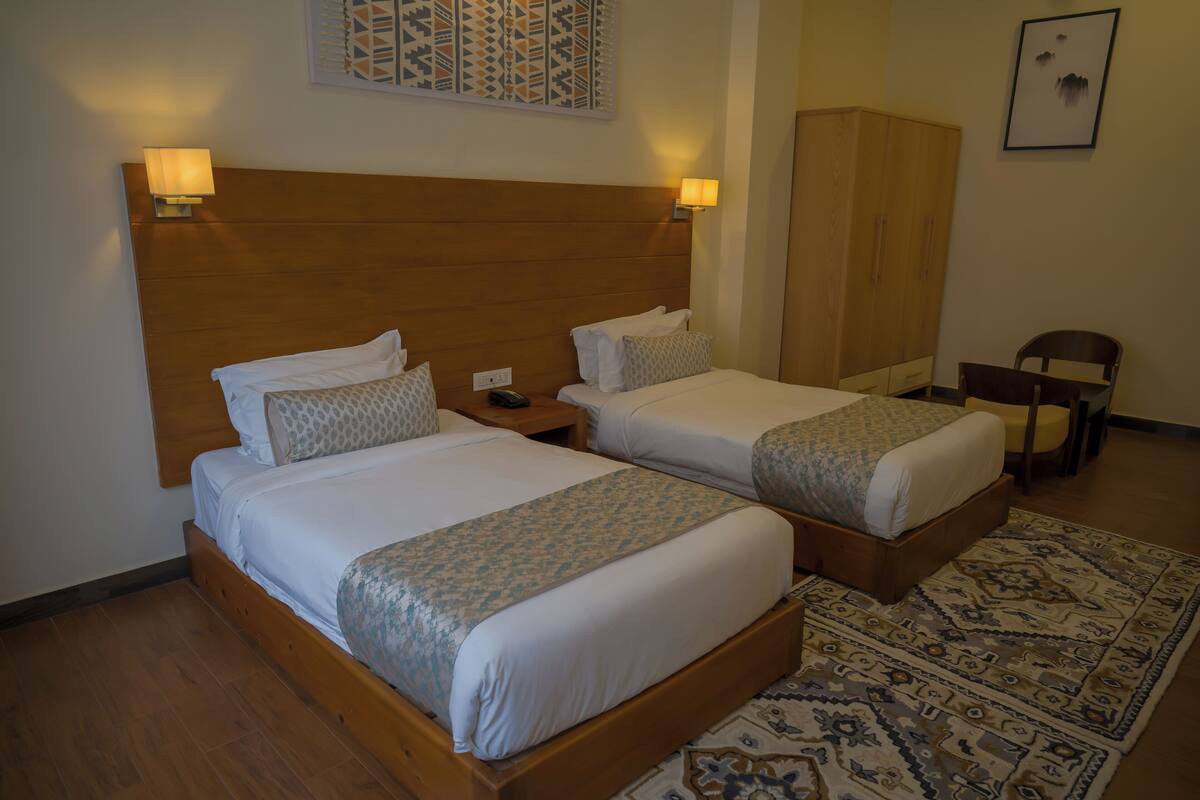 Une chambre d’hôtel avec deux lits simples, des têtes de lit en bois, des lampes de chevet, une armoire, une chaise et un tapis à motifs.
