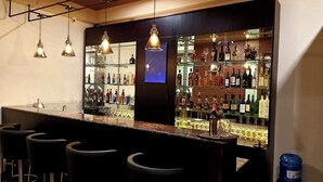 A bar area with a long counter, multiple stools, hanging pendant lights, and shelves stocked with bottles and glasses. - Legphel Hotel (Phuntsholing)