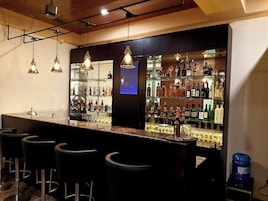 Una zona de bar con una barra larga, varios taburetes, lámparas colgantes y estantes llenos de botellas y vasos.