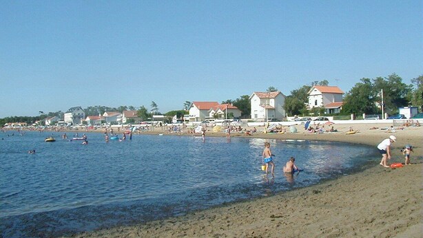 Een strand met mensen die zwemmen en staan in ondiep water, huizen op de achtergrond en een heldere lucht.