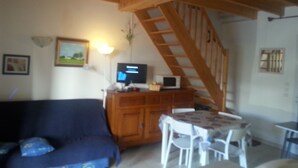 Een gezellige woonkamer met een houten plafond, een bank, een televisie, een eettafel met stoelen, en een trap.