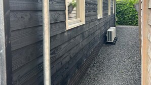 En byggnad med mörk träpanel, ett metallrör och en liten luftkonditioneringsenhet placerad på ett grusunderlag.