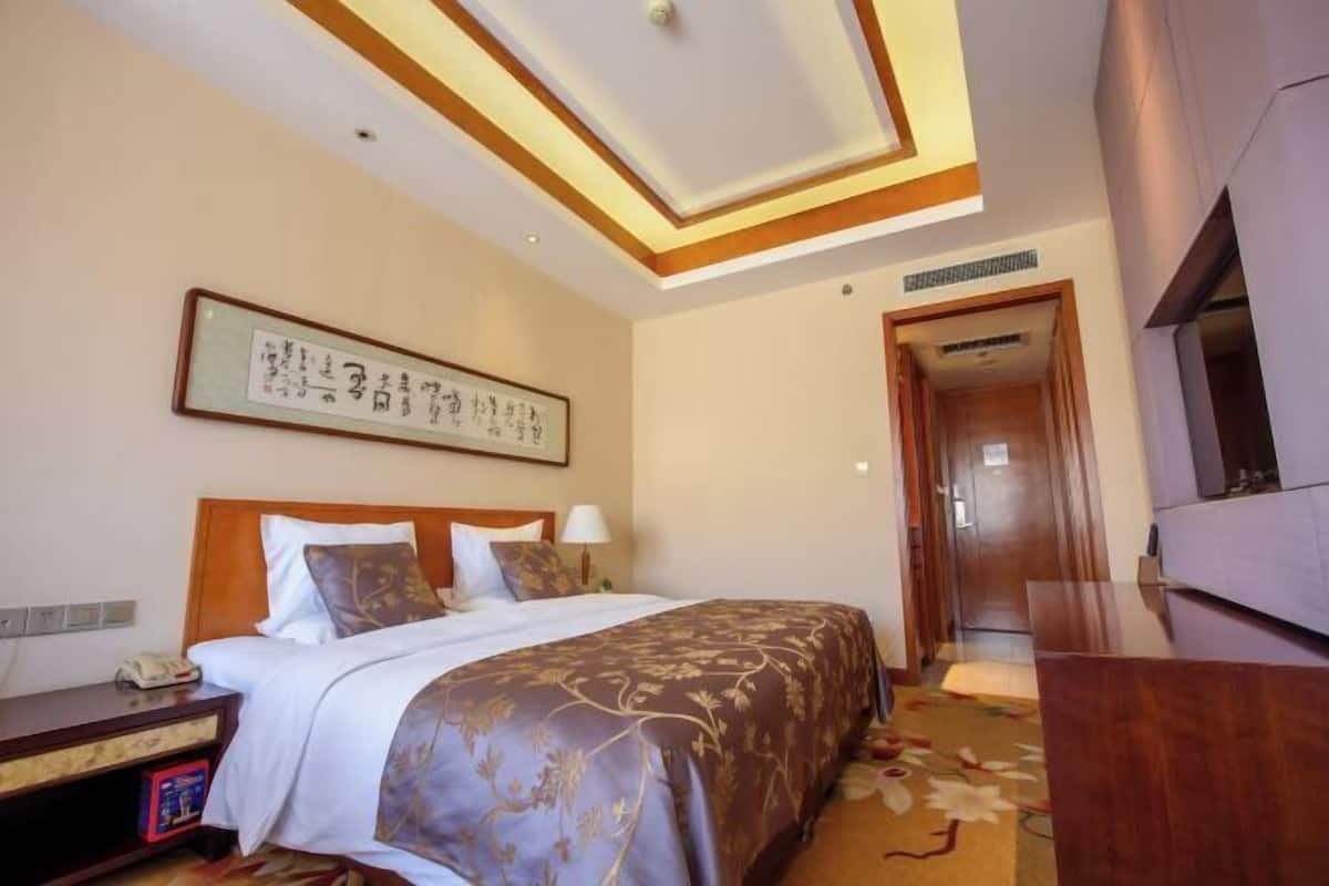 A hotel room with a neatly made bed, a bedside table with a lamp, a framed artwork on the wall, and a wooden wardrobe.